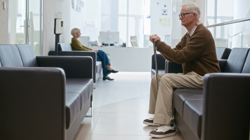 Long shot of aged Caucasian man sitting on sofa getting support from female doctor in white coat in hall of modern clinic