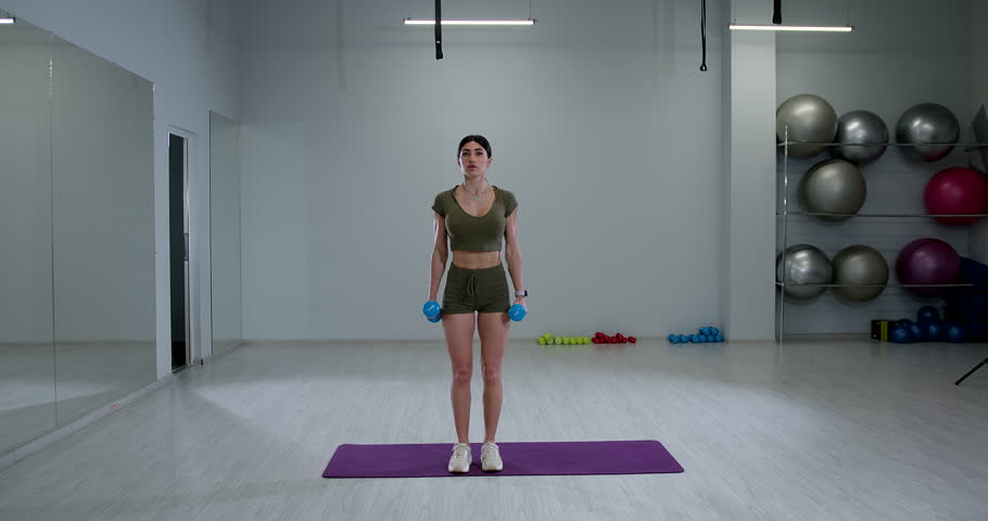 athletic woman is executing a standing one arm dumbbell bicep curl in a well lit gym. She appears focused and determined while working out.