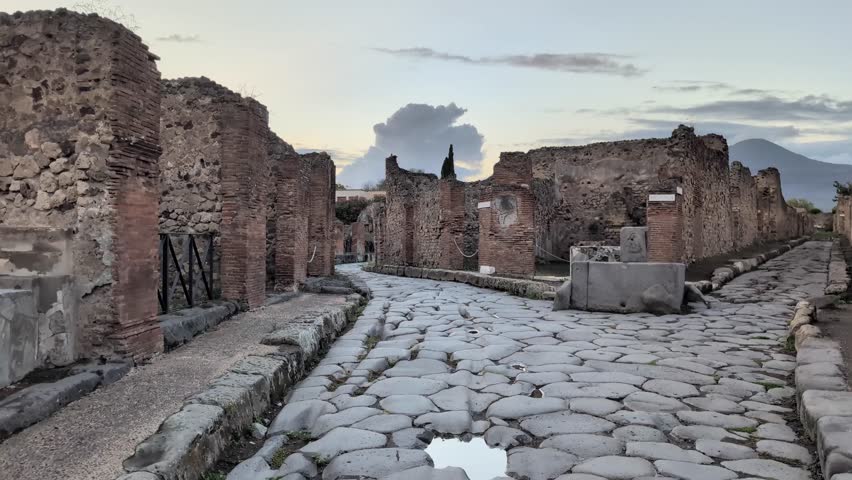 Slow pan of Pompeii’s ancient stone road glowing in golden sunset light, lined with ruins and framed by Mount Vesuvius in the distance. Perfect for travel or history footage.