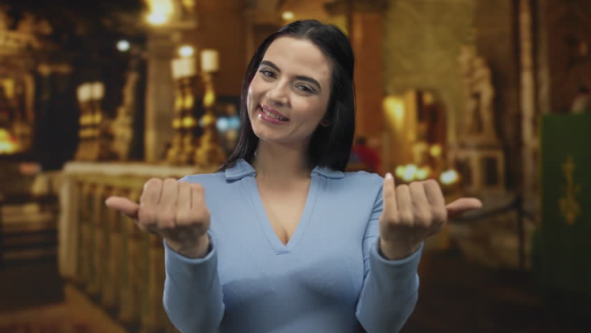 Woman gesturing in a church setting with inviting expression, wearing a blue blouse, conveying warmth and welcome indoors.
