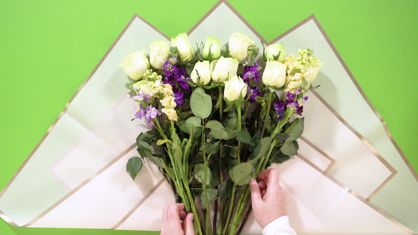 Hands place a full arrangement of white roses and multicolored stock flowers over layered pink-edged wrapping sheets on a green surface. The bouquet fills the frame.