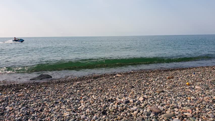 Sea waves hitting the shore of pebbles. Person riding jet ski. Outdoors
