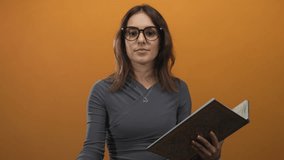 Young hispanic woman holding book blows kiss with hand to mouth in studio with orange backdrop; playful warmth. - Powered by Shutterstock - Get 15% off with code: PIKWIZARD15