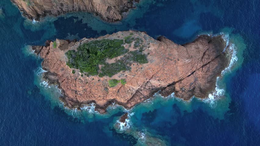 Cinematic aerial view of waves crashing against a small rocky island at the Mediterranean coast, with Esterel Mountains’ dramatic red cliffs in the background, French Riviera, Southern France – scenic