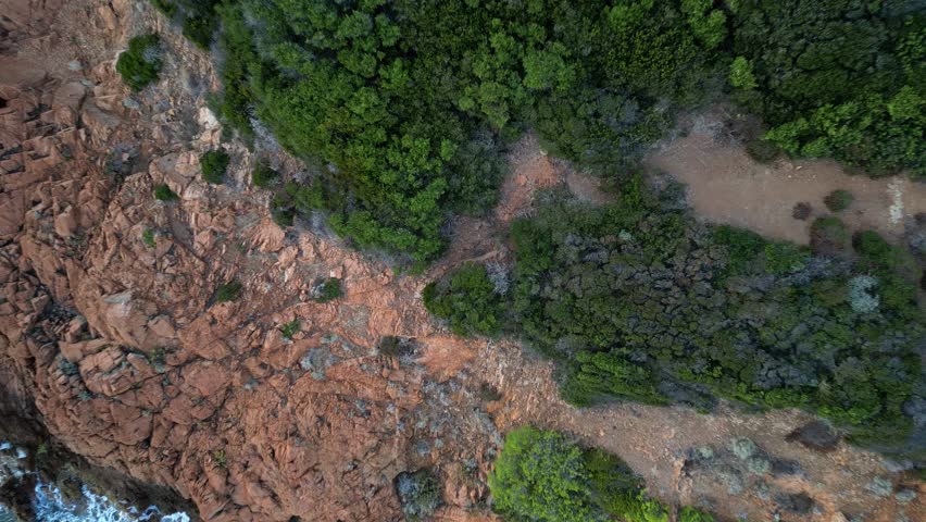 Cinematic aerial view of waves crashing against a small rocky island at the Mediterranean coast, with Esterel Mountains’ dramatic red cliffs in the background, French Riviera, Southern France – scenic
