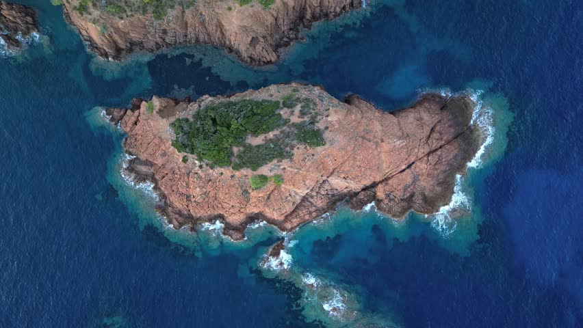 Cinematic aerial view of waves crashing against a small rocky island at the Mediterranean coast, with Esterel Mountains’ dramatic red cliffs in the background, French Riviera, Southern France – scenic
