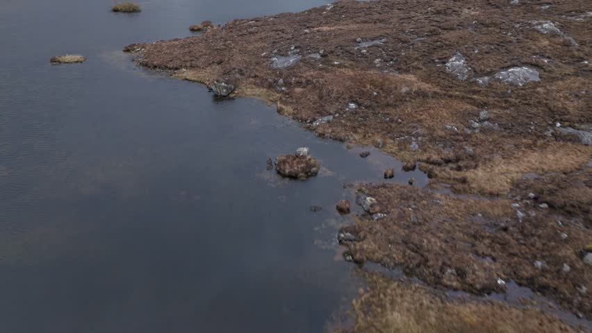 Cinematic drone opens low over an inland loch on the Isle of Harris, Scotland—tilts up while gliding forward to reveal rugged moorland and distant hills under moody grey skies.