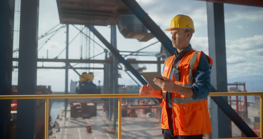 Male Logistics Coordinator in Safety Gear Shipment Manages Port Operations Using a Tablet, Optimizing Cargo Flow Cranes Loading Commercial Products Containersfor Shipping Processes.