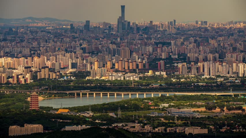 Aerial view of Beijing cityscape with modern skyscrapers, residential buildings, bridge over river, and mountains in background during golden hour lighting