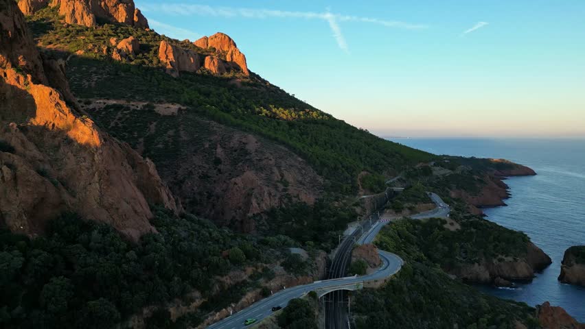 Aerial view of winding coastal road through Esterel Mountains at sunset, overlooking French Riviera cliffs, Mediterranean Sea, and scenic coastline near Saint Raphael, France
