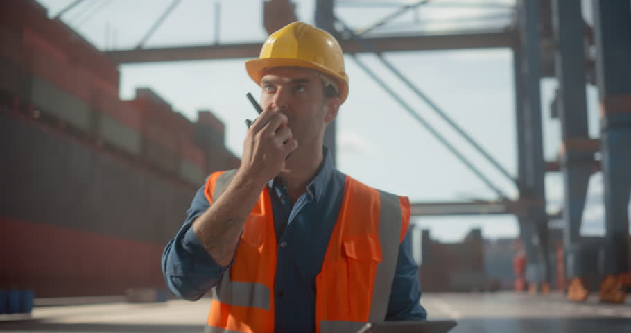 Male Logistics Manager in Hard Hat and Vest Shipment Coordinates Container Operations at a Busy Port Using a Tablet and Radio, Surrounded by Cranes and Cargo Containers.