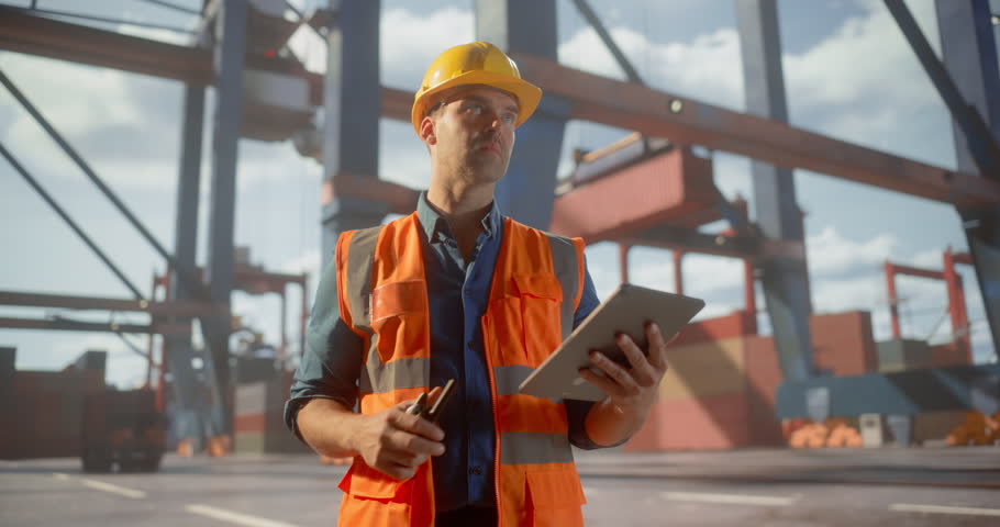 Male Logistics Manager in Safety Gear Uses a Tablet to Oversee Container Operations at a Busy Port, Optimizing Global Shipping Processes Cranes and Cargo.
