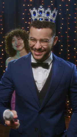 Vertical shot of smiling prom king, wearing crown and formal suit, giving speech and waving to crowd at high school party