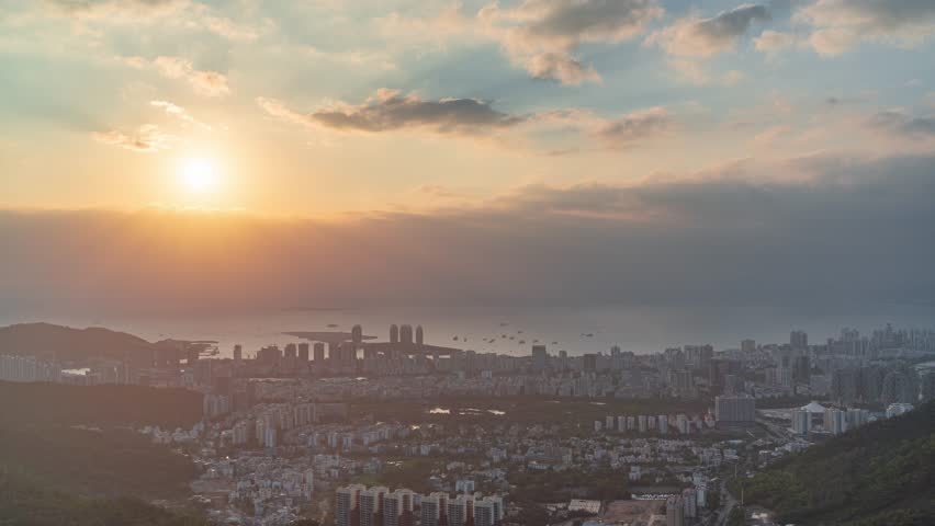 Stunning aerial view of Sanya city skyline at sunset with dramatic golden light illuminating the coastal urban landscape and surrounding tropical mountains.