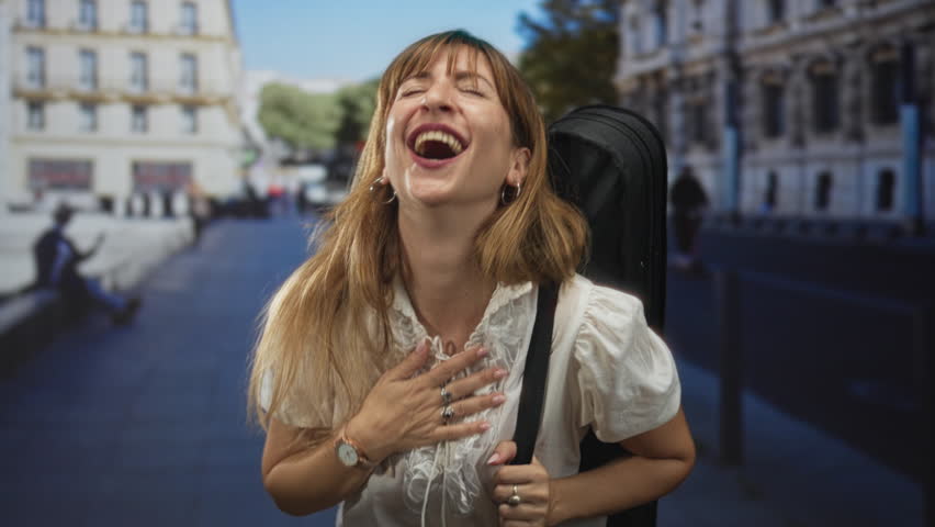 Woman laughing with hand on chest and gripping guitar case strap on a city street beside historic building, candid street scene; joy travel music.
