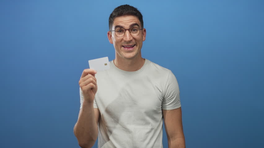 Man holding a white creditcard and cupping mouth while shouting in blue studio, wearing glasses and light tee; excitement promotion.