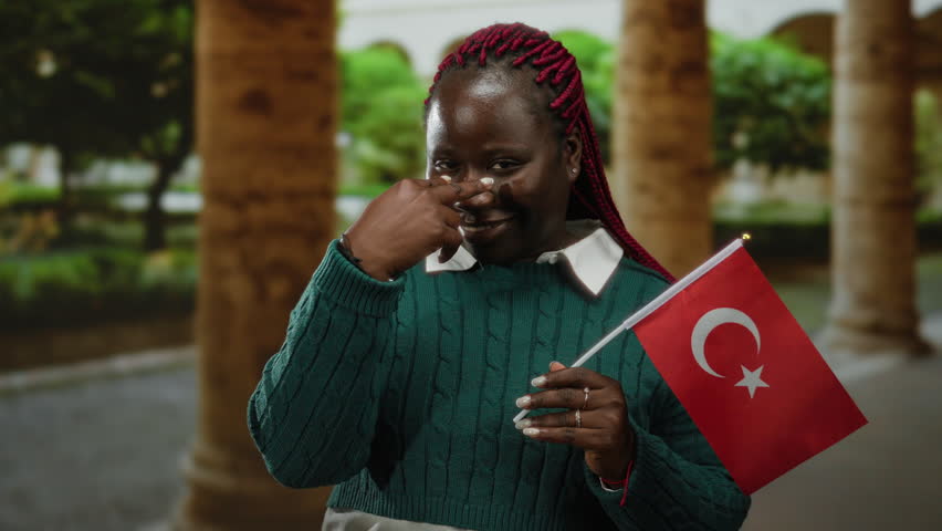 Woman smiling outdoors holding turkey flag in city street setting, showcasing culture with confident gesture in hand, autumn attire visible amidst architectural surroundings.