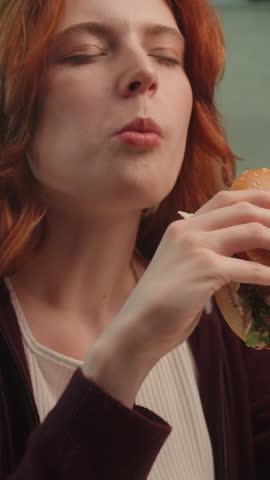 Satisfied Caucasian woman female girl eating junk fast food bite chewing tasty burger hamburger cheeseburger at home eat unhealthy meal diet nutrition disorder cheat meal overeating calories obesity