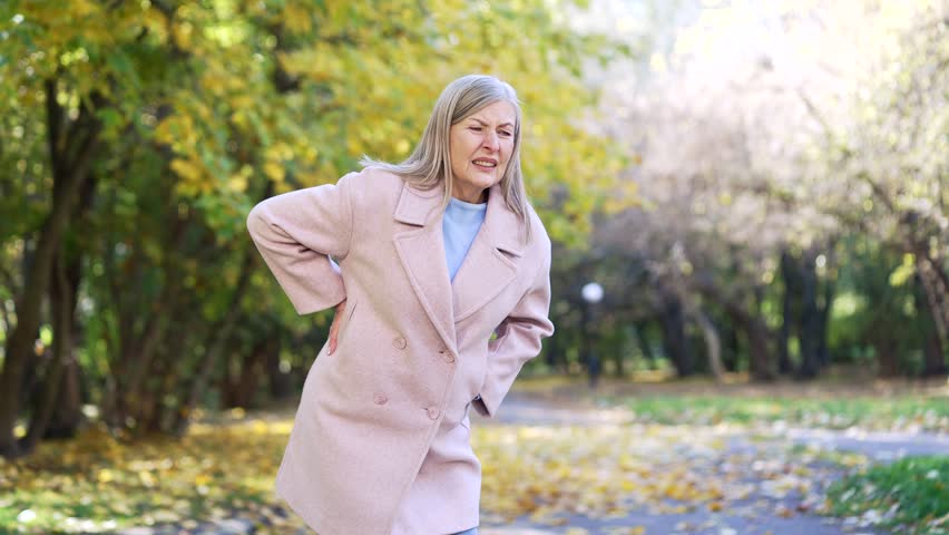 Senior woman feeling acute back pain outdoors. Eldery mature female holding spine with discomfort suffering from lower ache experiencing lumbar pain, painful posture in city ​​street outside