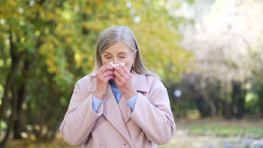 Sick cold senior woman in autumn park sneezing having headache. Elderly female outdoors suffering from flu symptoms, coughing, shivering, feeling weak, wrapped in warm coat, seasonal illness outside