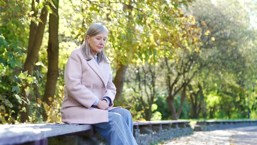 Elderly female with gray hair suffers from painful severe knee joint pain sitting on bench on street in urban city park. Upset woman massages leg muscles, could not walk, illness, cramps or rheumatism