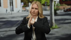 Young blonde woman on city street makes a fight gesture with a serious expression, surrounded by urban architecture and blurred scenery in the background. - Powered by Shutterstock - Get 15% off with code: PIKWIZARD15