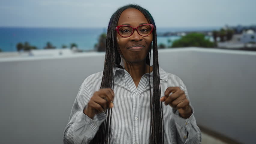 Woman with curly hair making air quotes on a seaside promenade wearing glasses against a beach backdrop with a relaxed expression under a cloudy sky.