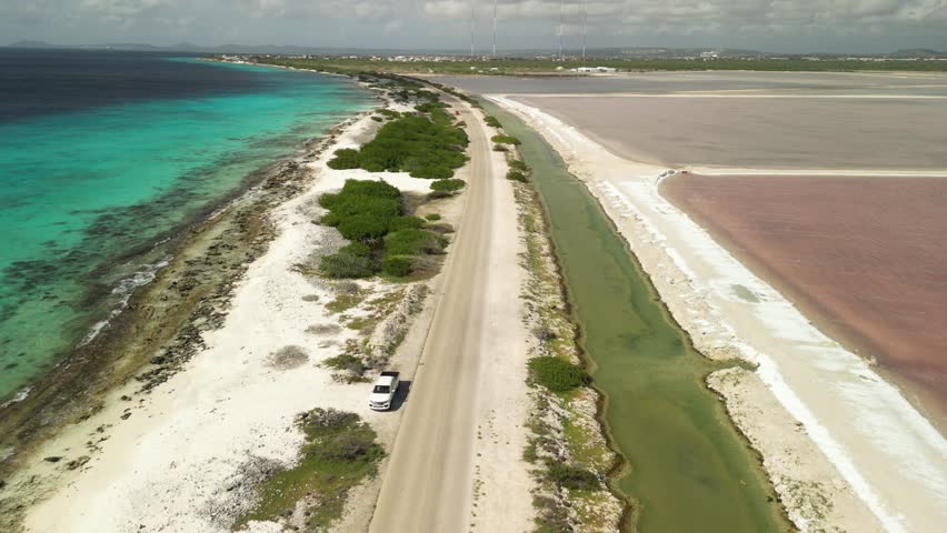 4K drone aerial of Bonaire’s pink salt lagoons beside the blue Caribbean Sea. Bright salt road runs between colours of pink, emerald, and turquoise — perfect for travel, nature, and cinematic visuals