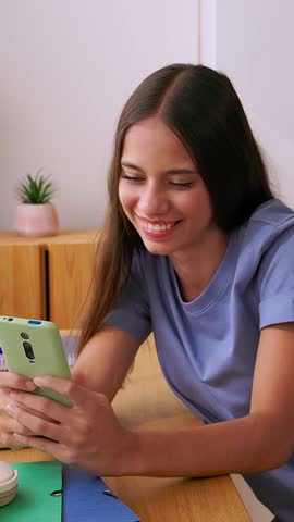 Vertical HD video of young smiling teenage student girl using mobile phone while while studying at her desk with a laptop. Social media, digital native people and communication lifestyle concept