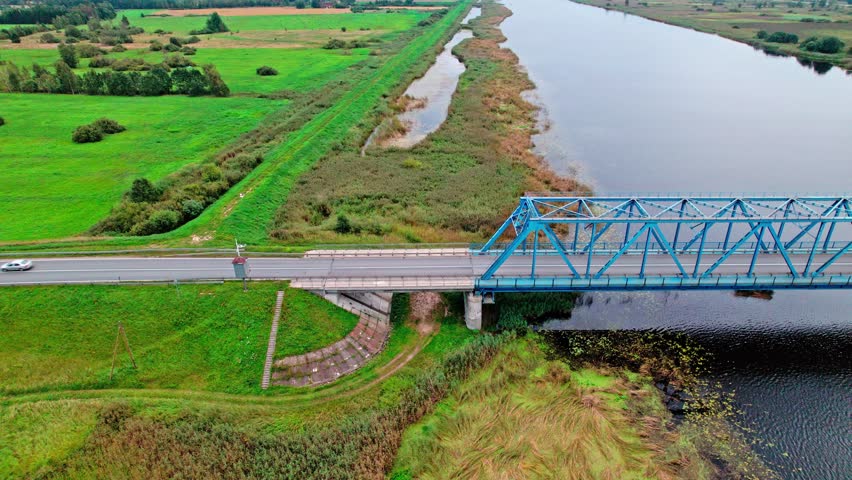 A stunning aerial view shows a blue metal bridge spanning a tranquil river surrounded by lush green fields in Latvia. Nature blends beautifully with the structure.