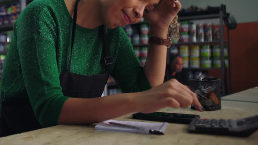 Female entrepreneur at a small business counter looking stressed, calculating finances with a notepad and calculator, facing business challenges in a retail setting