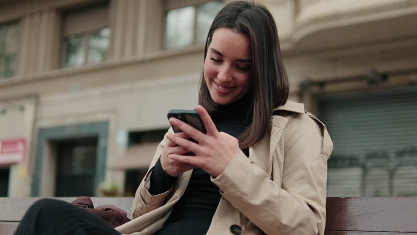 Smiling young woman using a smartphone while sitting on a bench. She is wearing a black turtleneck and beige trench coat.