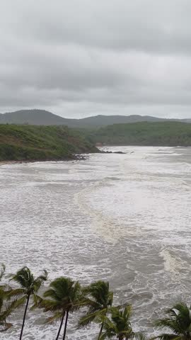 Dramatic overhead view of a tropical beach during high tide or a storm, showing powerful, churning white waves rolling onto the shore beneath lush green, mist-covered hills. 