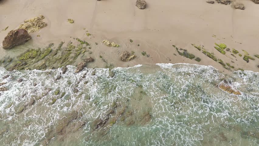 Low drone shot of soft sea waves washing over rocks and sand on a turquoise beach, forming natural white foam patterns on a calm bright day.