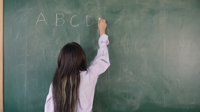 Video English basic class online concept. Happy young Asian female teacher stands writing and teaching English language lesson on the blackboard via video call on a technology smartphone in classroom.