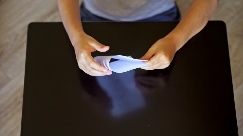 A 9-year-old boy sits at a table and folds a paper airplane from a white sheet, top view, closer than medium shot with selective focus on hands.
