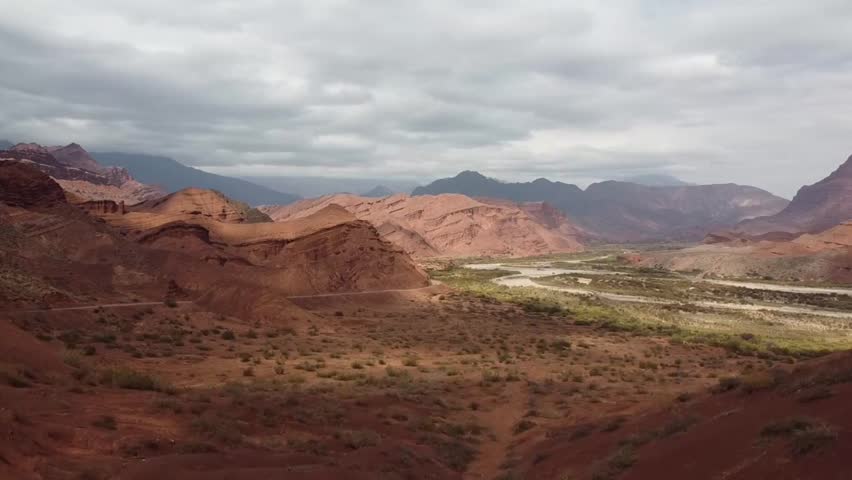 Drone flying over the red and pink canyon near Cafayate, Argentina, with a river running through the valley and a clear blue sky above. The wide panoramic landscape captures the natural beauty