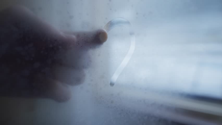Close up on glass window as hand draws heart shape with finger on cold day. Condensation fog on apartment window house in winter season day. Man marks heart in romantic gesture in freezing weather