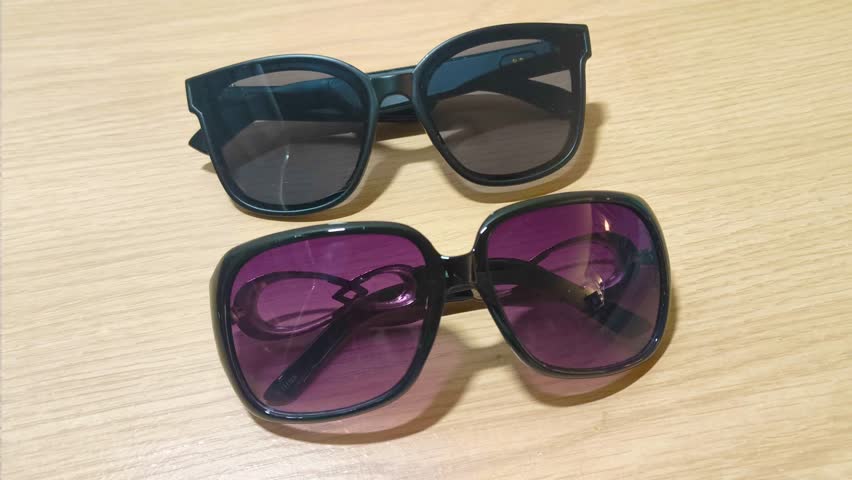 Two stylish pairs of sunglasses, one with dark lenses and the other with vibrant purple lenses, rest on a light wooden surface, hinting at summer fashion and outdoor activities.