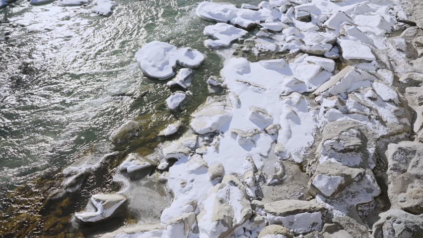 Freezing cold water flowing in small town or village river or stream with snow. White snow pieces between rocks on fresh cold water from European Alps. Hot weather end of winter melting down snow