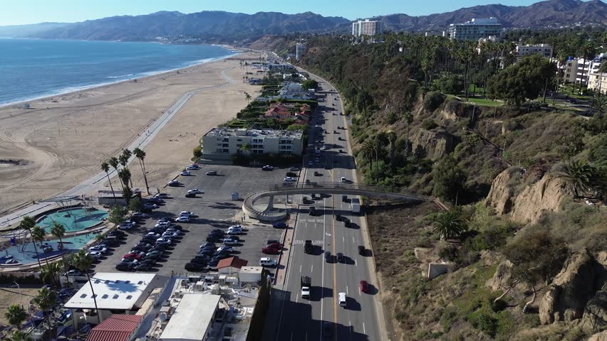 4K aerial drone footage of Santa Monica, California, showing Pacific Coast Highway with moving cars along the coastline. The scenic road connects the Pacific Ocean, sandy beach, and the Santa Monica M