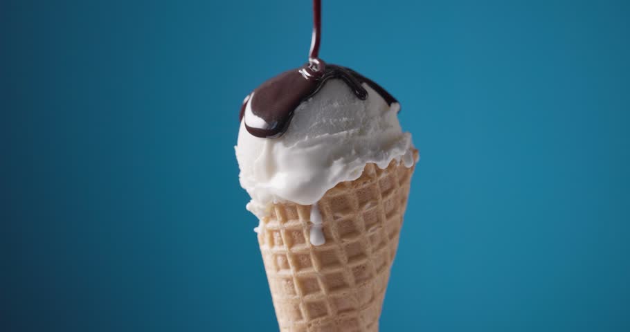 Melted chocolate sauce flowing over a scoop of vanilla ice cream in a waffle cone, set against a blue background
