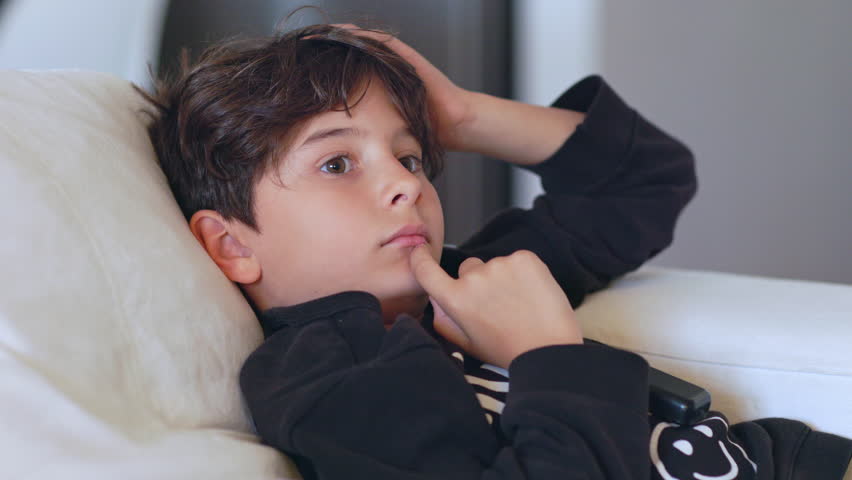 Close-up shot of an 8-year-old boy sitting on a sofa, eyes wide and mouth slightly open as he watches television, completely captivated while holding the remote in his hand.	