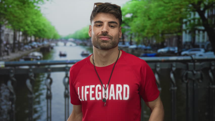 Lifeguard man wearing red shirt gestures open palms beside canal railing on street with sunglasses perched on head; confidence.