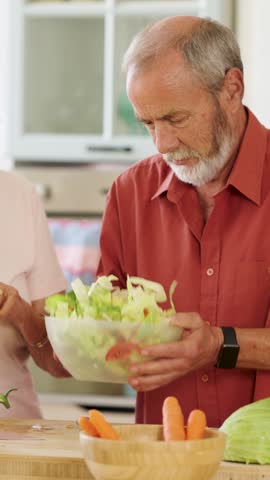 Elderly man preparing a healthy salad with the help of his wife, adding spices and mixing ingredients in a bowl in their kitchen. Vertical footage