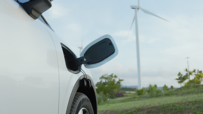 Woman plugging electric car charging cable in countryside with wind turbine background, renewable energy
