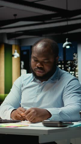 Vertical Video Black office employee ripping a piece of paper after making a mistake, restarting his task in anger and tearing up the page sheet. Discontent guy writes a typo while transcribing
