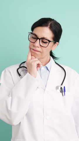 Pensive middle-aged female doctor wearing a lab coat and stethoscope thinking and then finding a brilliant solution on a turquoise background. Vertical footage