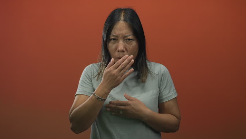 Woman covering mouth with hand and holding chest in orange studio with plain backdrop, wearing casual tshirt and bracelet, young latina; nausea.
