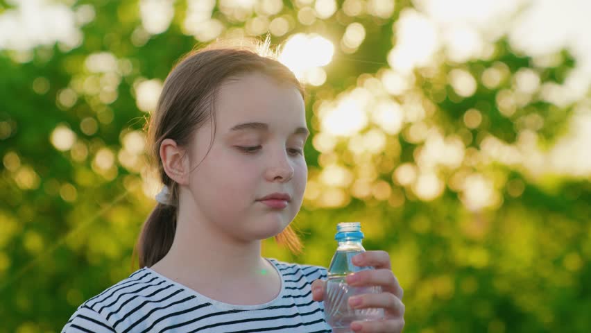 Child girl drinks refreshing water from bottle, walk, park, nature. Beautiful girl drinks clean mineral water outdoors, summer heat in park. Exercise outdoors, quench your thirst with cool water.